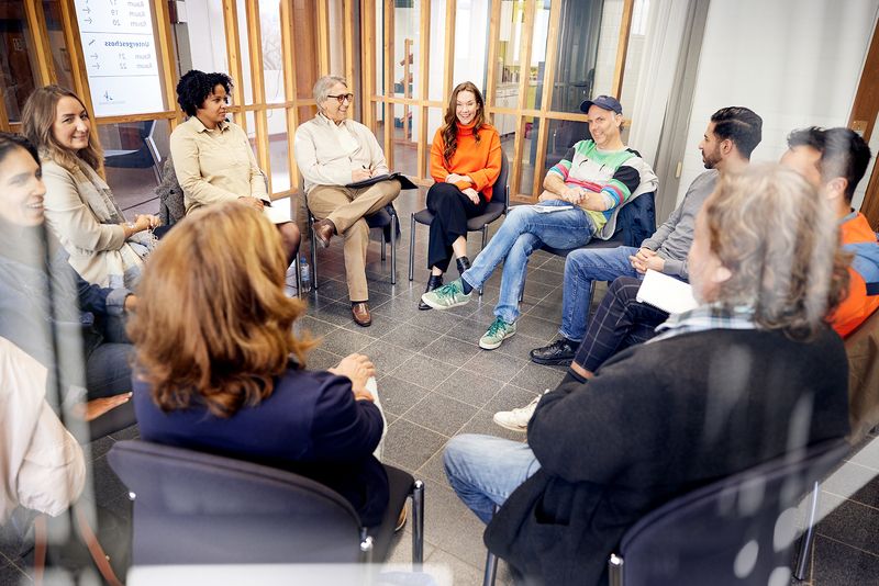 Zehn Menschen sitzen in einem Stuhlkreis zusammen. Sie besprechen etwas. Einige Personen sind mit Gesicht zu sehen, andere nur von hinten.