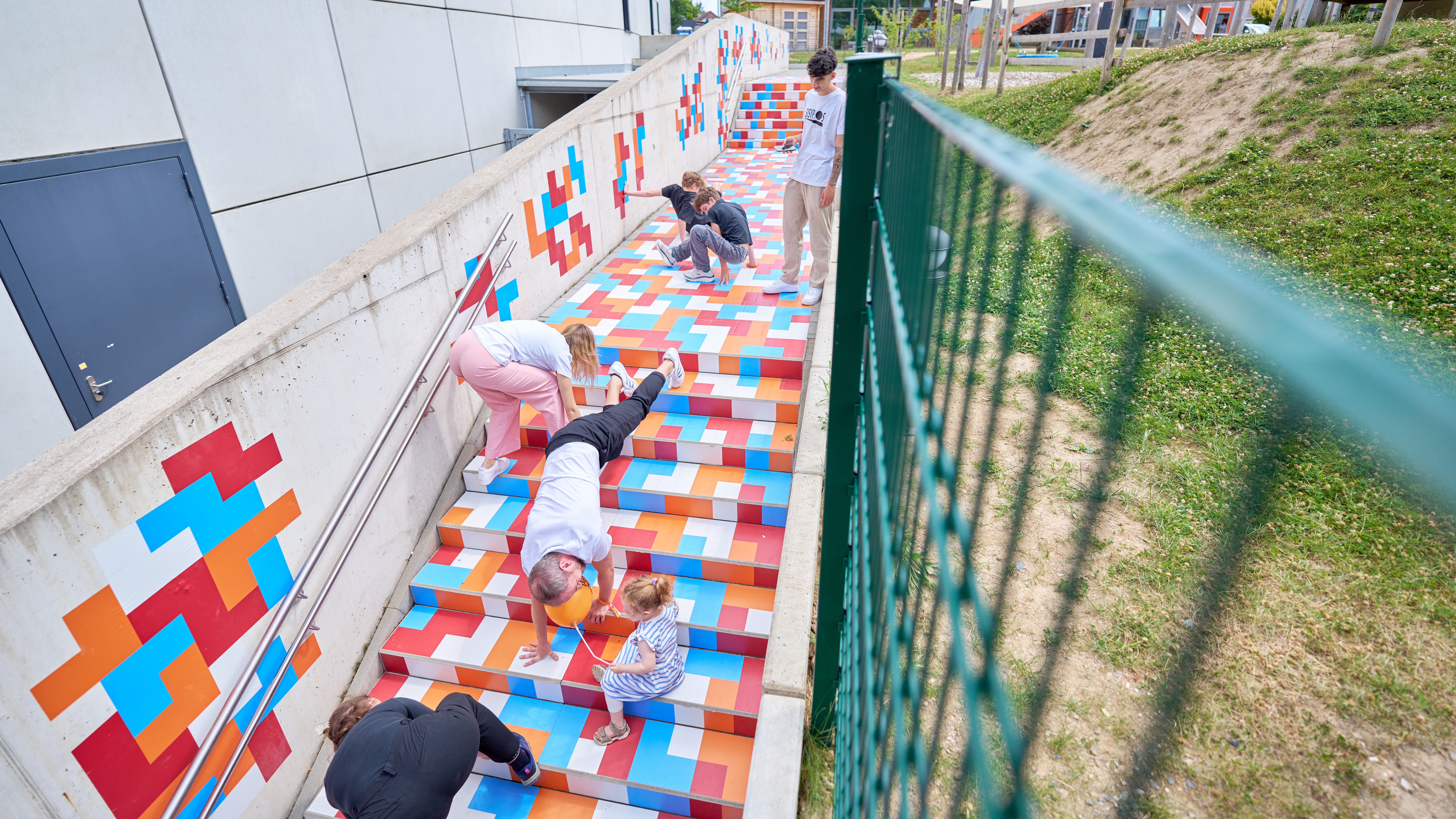 Eine mit bunten Rechtecken beklebte Treppe. Auf der Treppe sind Menschen, die mit Händen und Füßen auf verschiedenen Farbrechtecken stehen.