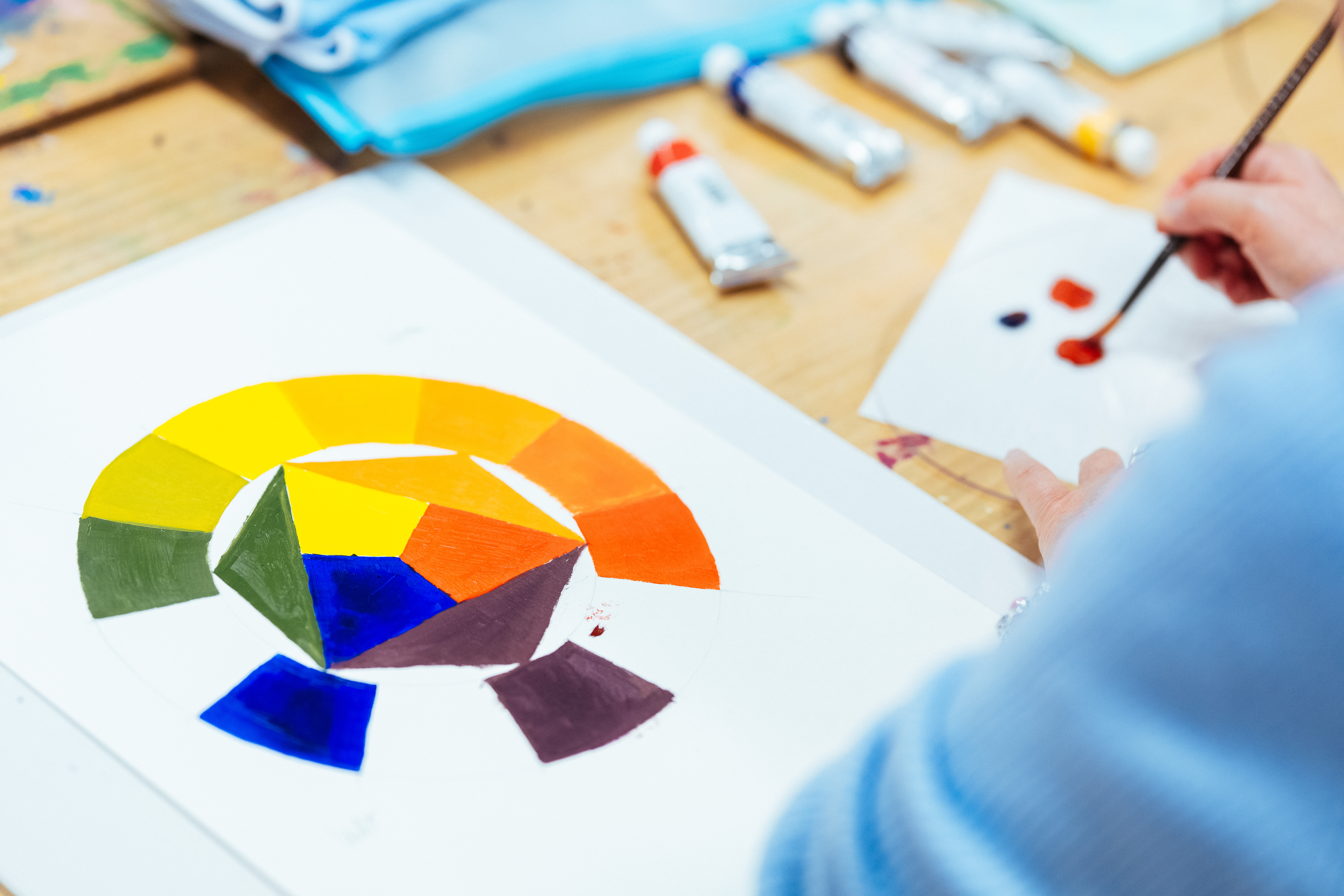 Auf einem Tisch liegt ein Papier, auf das ein Farbkreis gemalt wird. Am Bildrand unscharf ist eine Hand, die einen Pinsel hält, der gerade in Farbe getunkt wird.