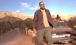 Ein Mann steht in einer Wüste vor einem schrottreifen Auto. 