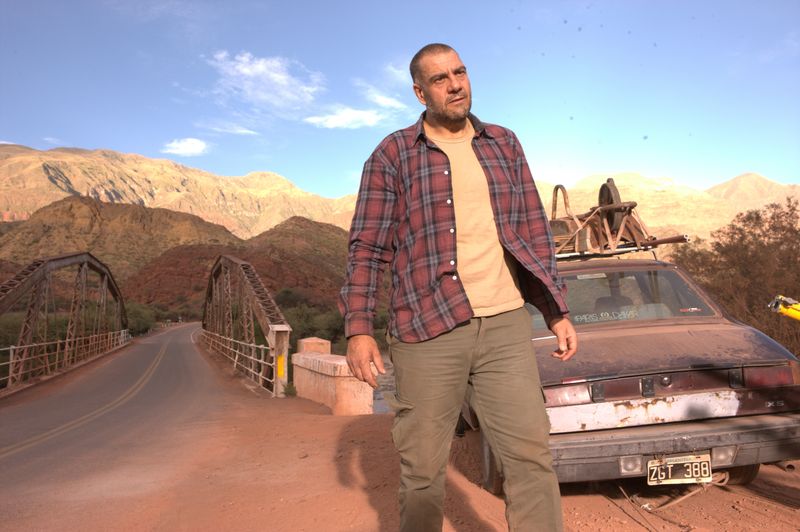 Ein Mann steht in einer Wüste vor einem schrottreifen Auto. 