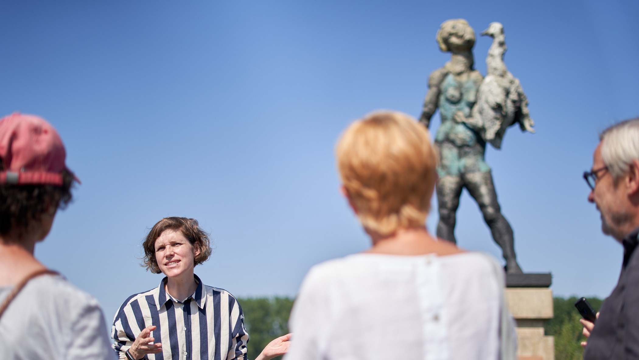 Mehrere Menschen vor einem Kunstwerk im öffentliche Raum. Eine Person erzählt etwas, die anderen hören ihr zu.