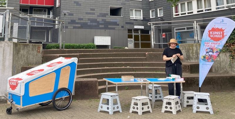 Im Stadtraum aufgebaut ein Tisch, mehrere Hocker und das Kunstschulmobil. Eine Person, die eine Aktion vorbereitet.