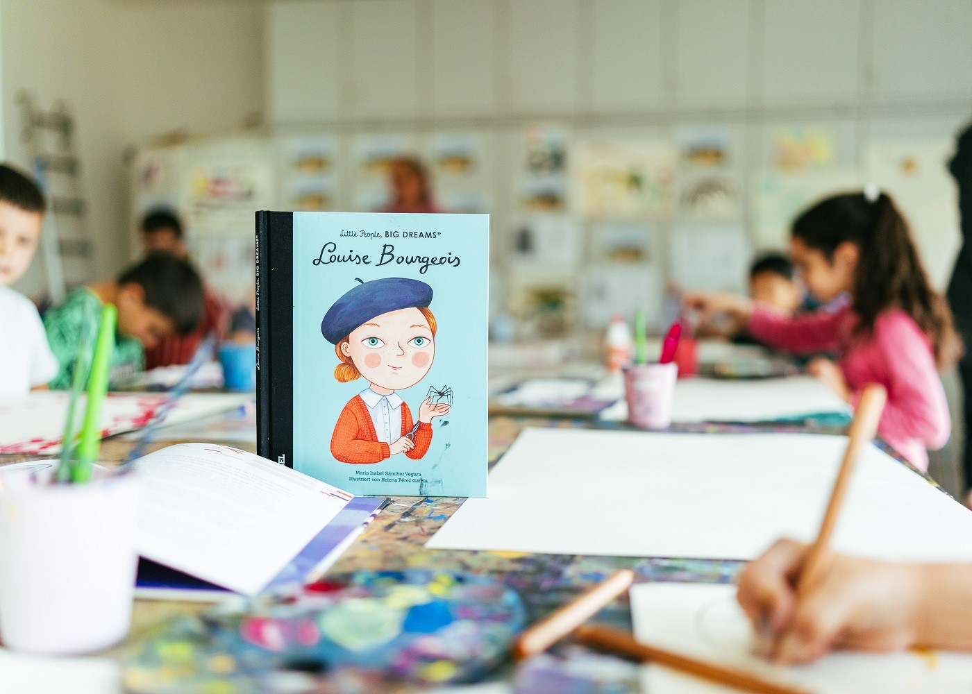 Blick auf einen Tisch mit Papieren, Stiften und Materialien. An dem Tisch sitzen Kinder. Auf dem Tisch mittig steht ein Buch zu Louise Bourgeois.