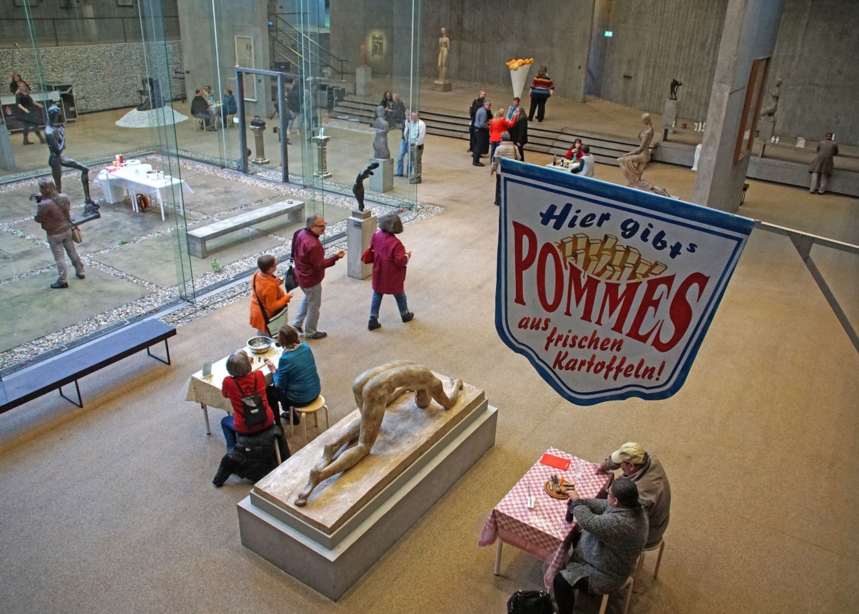 Blick von oben in eine Ausstellungshalle. Mittig ist eine Statue, daneben zwei Tische mit zwei Personen. Mittig hängt ein Schild mit der Aufschrift 