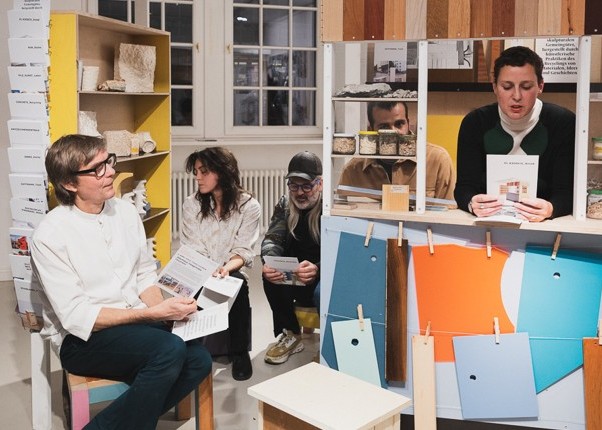 Mehrere Menschen in einem Raum, sitzend. An einer Wand steht ein volles Bücherregal. Rechts steht eine Theke, auf die sich eine Person stützt und etwas vorliest.