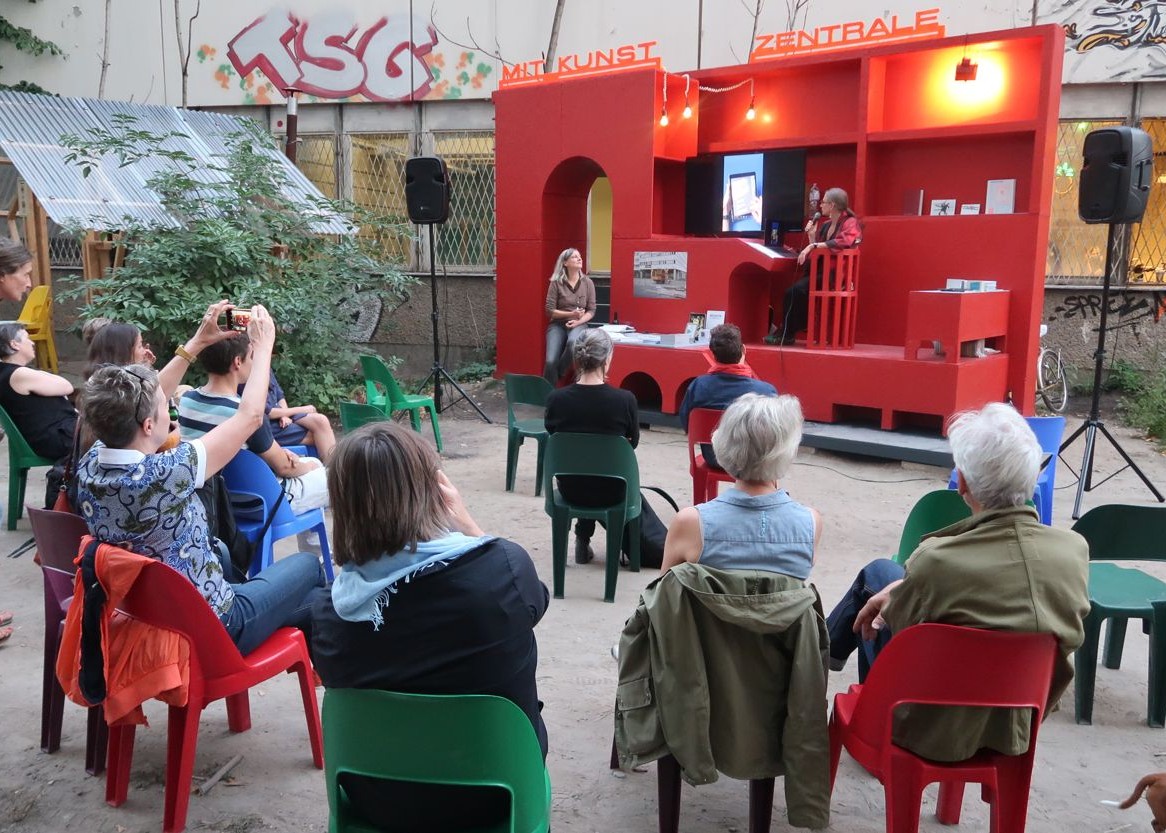 Mehrere Menschen sitzen in einem Stuhlkreis. Vor ihnen ist, der Kamera zugewandt, ein rotes, großes, regalähnliches Gebilde, in dem Bücher stehen und vor dem die Künstlerinnen der Mitkunstzentrale sitzen.