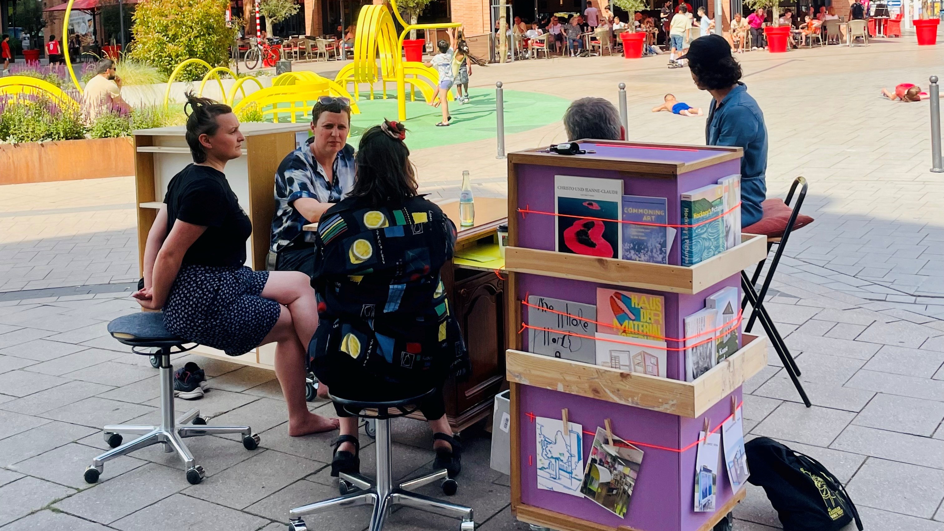 Mehrere Menschen sitzen im Stadtraum zusammen. Vor ihnen ein Stand mit Infomaterialien.