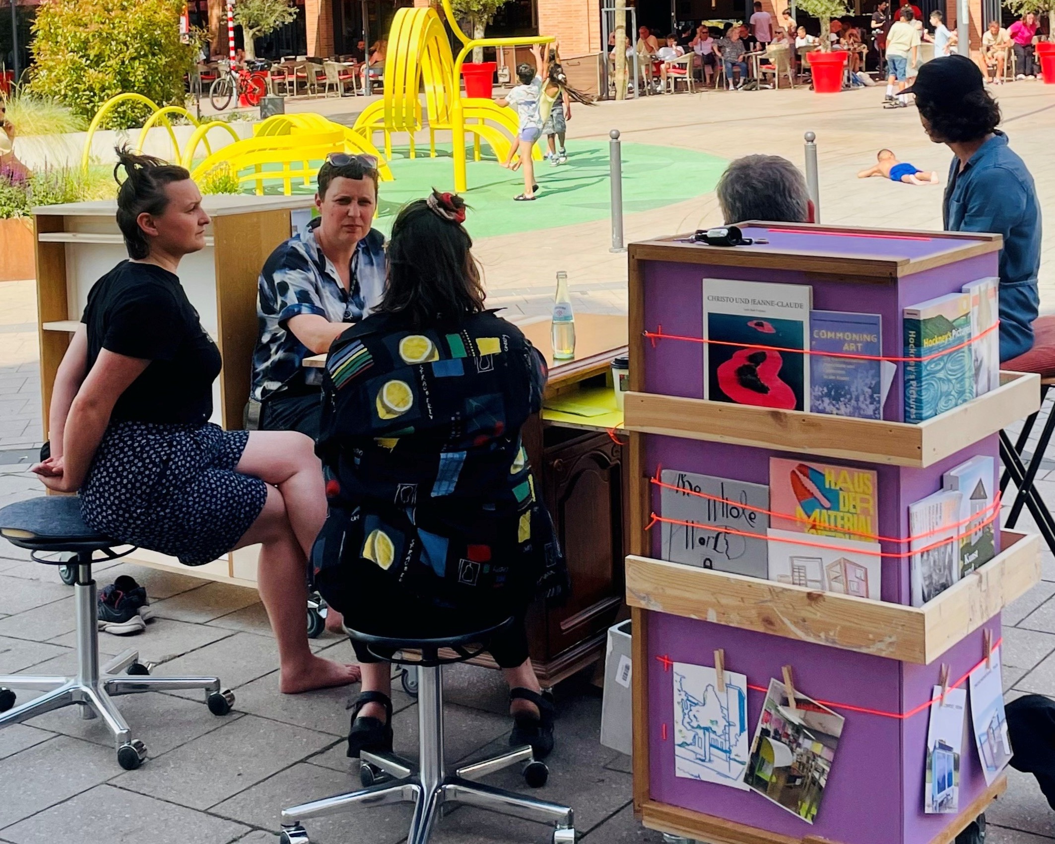Mehrere Menschen sitzen im Stadtraum zusammen. Vor ihnen ein Stand mit Infomaterialien.