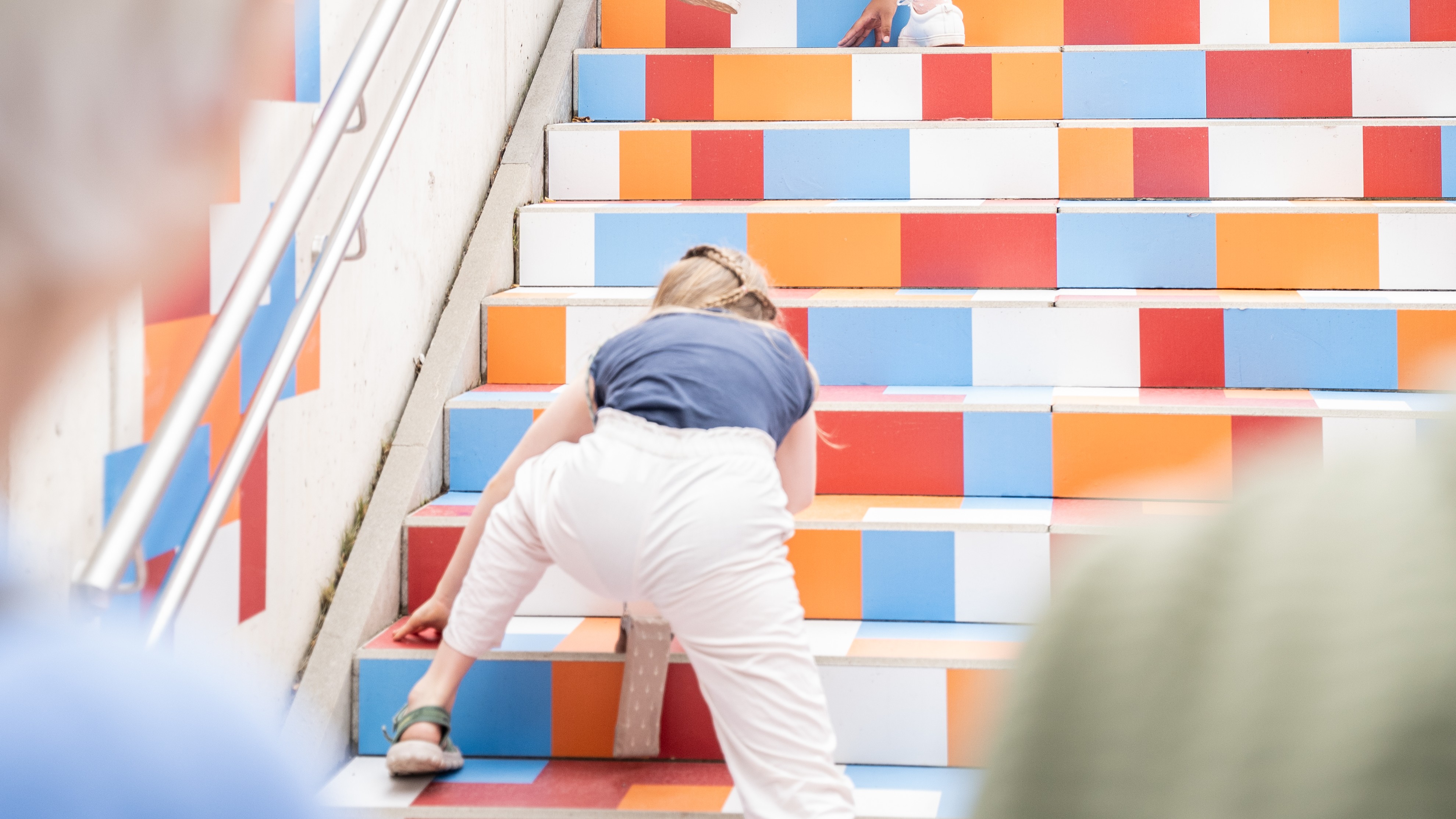 Eine Person, die eine mit bunten Rechtecken beklebte Treppe hochklettert, indem sie die Hände und Füße auf unterschiedlich farbigen Flächen hat.